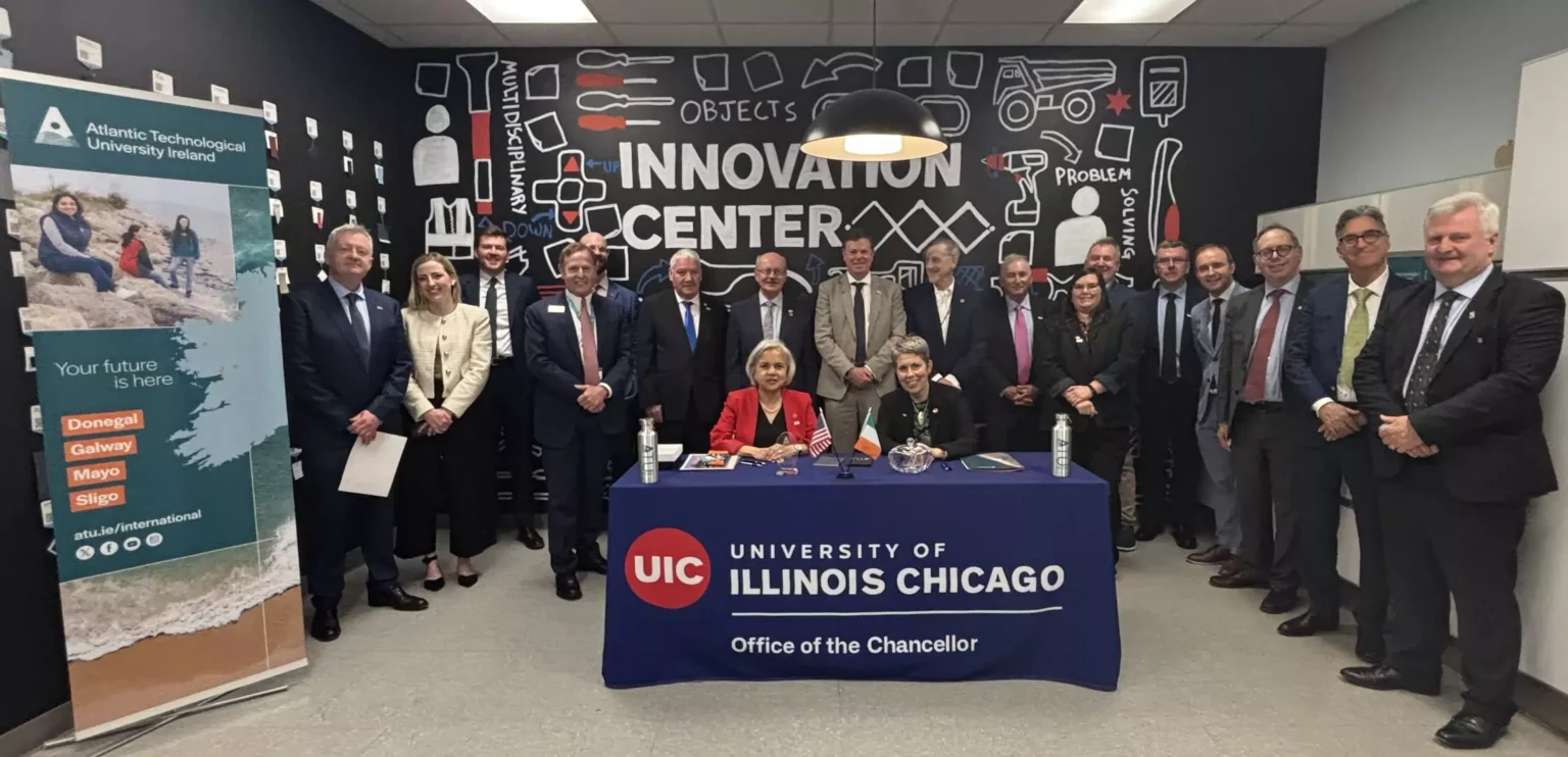 A group of peple including officals and representatives from Atlantic Technological University Ireland and University of Illinois Chicago, pose for a photo in the UIC Innovation Center. Two women are seated at a table with the UIC logo and "Office of the Chancellor" text, likely signing a partnership or agreement. A promotional banner for ATU Ireland is displayed on the left, and a wall with innovation themed illustrations is in the background.