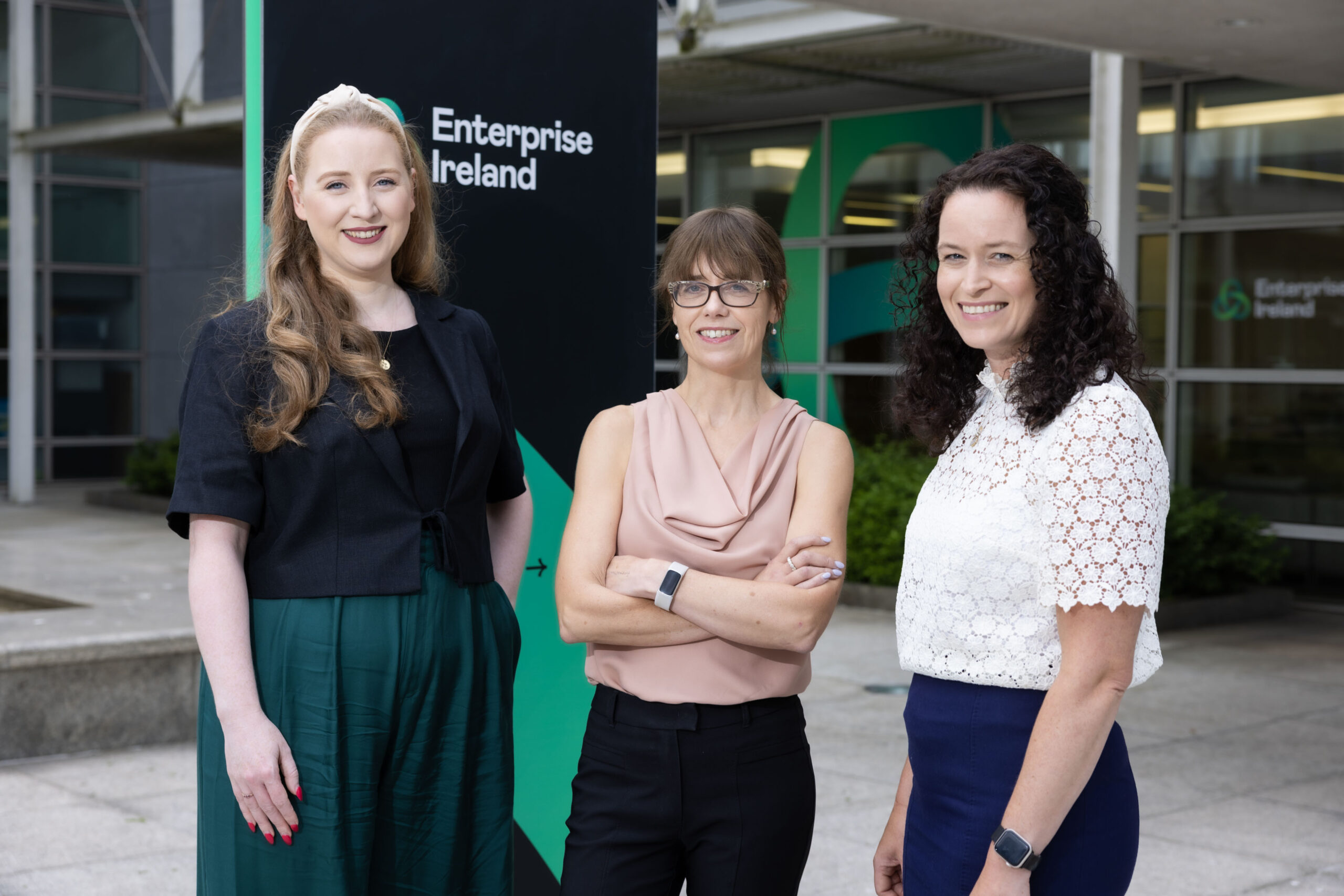 EIC-accelerator-pic-scaled Three women standing in front of an Enterprise Ireland building. They are smiling at the camera. One woman wears a dark jacket over a black top and green skirt, the second wears glasses and a sleeveless blush top, and the third wears a white lace blouse with a navy skirt.