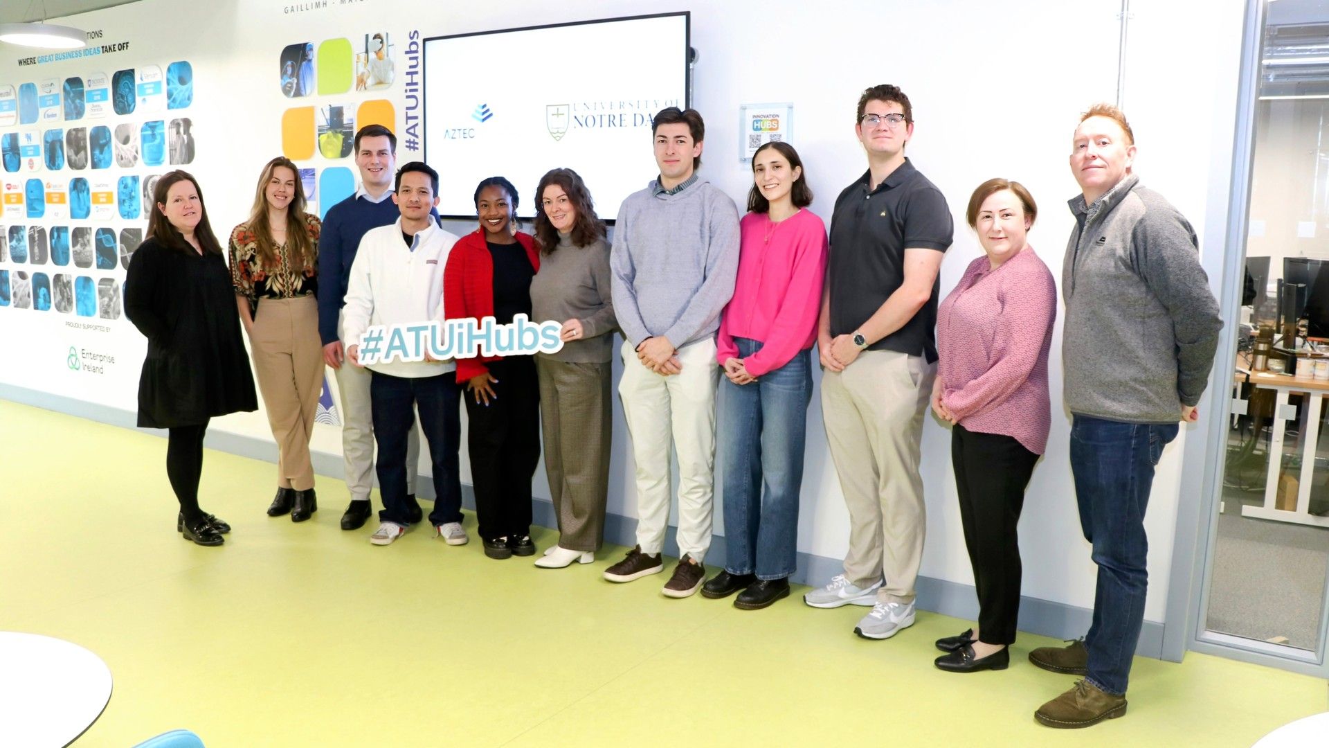 Three women standing in front of an Enterprise Ireland building. They are smiling at the camera. One woman wears a dark jacket over a black top and green skirt, the second wears glasses and a sleeveless blush top, and the third wears a white lace blouse with a navy skirt.